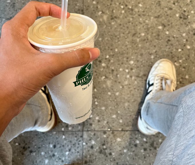 A hand holds a Phuc Long bubble tea cup with the text "PHUC" and "www.phuclong..." visible on it, while the person stands on a speckled floor wearing jeans and sneakers. The image humorously symbolizes the "Tổng chi phí 1 năm tập làm Trader lướt sóng" (total cost of one year of learning to day-trade), with the overpriced iced drink representing the hidden, everyday expenses that drain a beginner trader's budget — much like the unspoken fees and small costs that quietly accumulate alongside the major losses.

Caption: "Just like this Phuc Long drink — small daily purchases that quietly drain a beginner trader's wallet, on top of the 30M burned in Futures and 12M lost in fees."