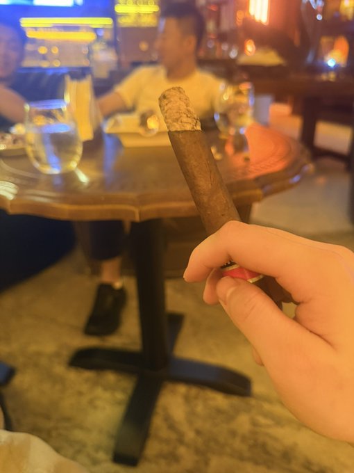 A hand holding a lit cigar in the foreground with blurred people seated at a small wooden table in a dimly lit lounge or bar setting.