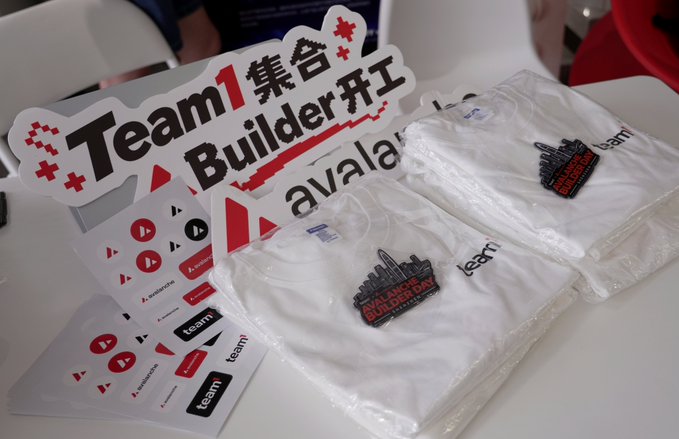A team-building event display with a "Team 1 Builder 集合开工" sign, Avalanche-branded stickers, and folded white t-shirts in plastic packaging.