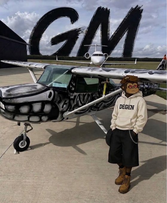 A person wearing a hooded sweatshirt reading 'DEGEN', black shorts, sunglasses, and a pilot's cap stands in front of a small airplane painted with a skull design, with 'GM' spray-painted on the side of the building behind it.