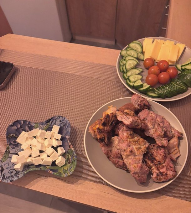 A meal spread on a wooden table featuring grilled meat, a plate of sliced cucumbers, cherry tomatoes, cheese, and a decorative bowl of cubed cheese.