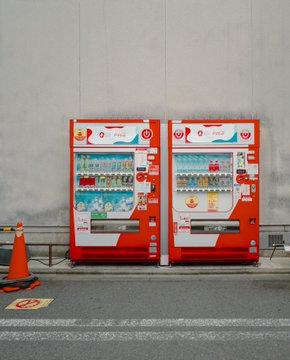 The image caption should focus on describing the actual visual content - the two vending machines - rather than the project's promotional message.

Two vibrant red and white vending machines stand side-by-side against a plain concrete wall, stocked with an assortment of bottled drinks, while an orange traffic cone with a black-and-orange striped base sits on the sidewalk nearby.