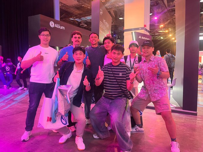 A group of people posing together at what appears to be a gaming or tech convention, with booths for "Allium" visible in the background.