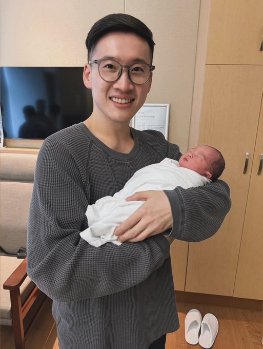 A smiling young man with glasses holding a newborn baby wrapped in a white blanket in what appears to be a hospital room.