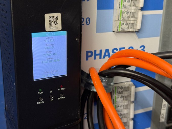 A close-up photograph of an electrical monitoring device's LCD screen displaying readings (Current 91.81A, Voltage 236.9V, Power 21692.0W, Energy 13558.6kWh) mounted near orange cables and circuit breakers labeled 'PHASE 2'.
