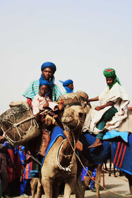 Several people wearing traditional Tuareg blue and green headwraps and robes ride camels loaded with cargo containers across a desert landscape.