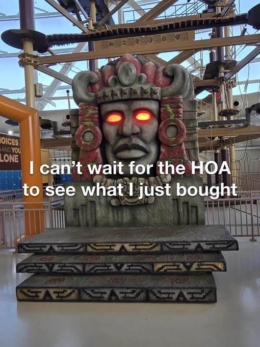 A large stone statue resembling an Aztec or Mayan deity with glowing red eyes stands inside an indoor amusement park ride area, accompanied by the caption "I can't wait for the HOA to see what I just bought."