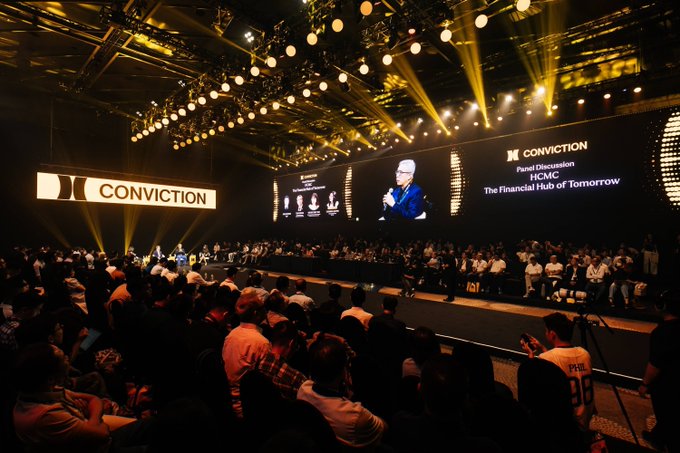 A large audience fills a dimly lit conference hall at the Conviction event, where a speaker is shown on a giant screen delivering a "Panel Discussion: HCMC – The Financial Hub of Tomorrow," alongside the "CONVICTION" branding displayed prominently on stage. The scene captures the scale and atmosphere of the crypto, blockchain, and AI gathering taking place in Ho Chi Minh City.