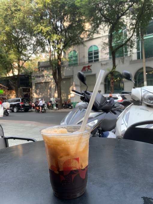 The image shows a table with an ice cream drink sitting on top of it, which appears to be a milkshake or smoothie. The drink has a straw sticking out of it. In the background, there are several motorcycles parked outside a building, including one motorcycle that is closer to the foreground and another further back in the scene.