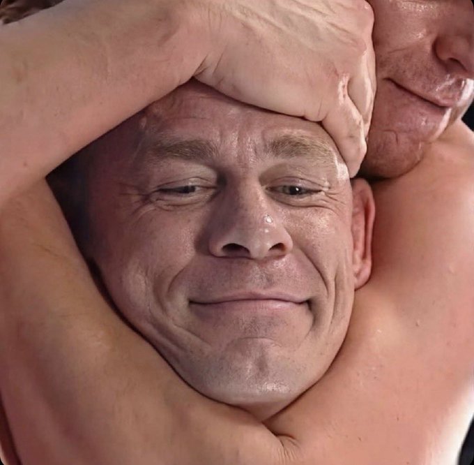 A close-up portrait of a man with light hair and blue eyes, smiling slightly while his head is surrounded by the muscular, bare arms of others.