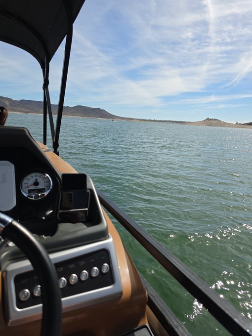 A view from a boat showing the dashboard with steering wheel, speedometer, and control panel, with green lake water and distant mountains under a blue sky.