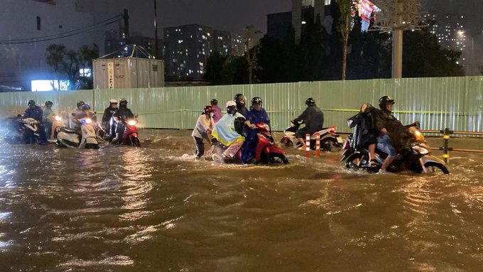 A nighttime photograph showing several people riding motorcycles through a heavily waterlogged street or flooded road, with buildings and a billboard visible in the background.