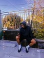 A small, low-resolution image shows a person wearing a dark hooded sweatshirt and shorts squatting on a tiled rooftop or balcony with trees in the background.