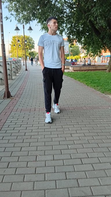 A young man walking on a brick sidewalk in casual clothing, wearing a light blue t-shirt, black cargo pants, and sneakers.