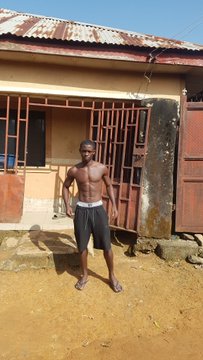 A shirtless young man in dark shorts stands barefoot on a dirt path in front of a small building with a rusty corrugated metal roof and red-framed open doors.