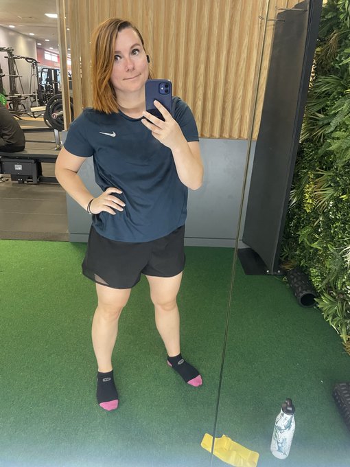 A woman in athletic wear taking a mirror selfie at a gym, wearing a navy Nike shirt, black shorts, and distinctive black socks with pink and purple toes.