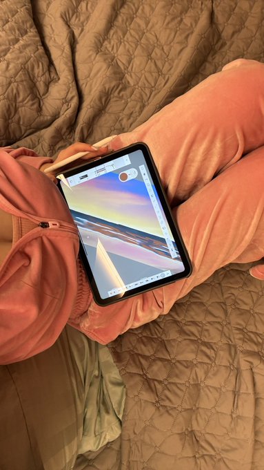 A first-person view of a person wearing pink sweatpants sitting on a quilted bed, holding a tablet that displays a scenic landscape image with a sunset over water.