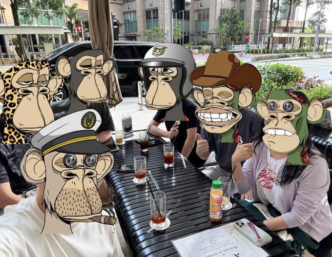 The image features a group of six anthropomorphic monkeys sitting around a dining table outdoors. The monkeys are wearing hats and masks, adding a whimsical touch to the scene. There are also people present at the gathering, with one person seated on the left side of the table and another person standing near the right edge of the frame.

The table is set with various items such as cups, bottles, and a book, suggesting that they might be enjoying some refreshments or engaging in conversation while sharing stories about their travels. The overall atmosphere appears to be relaxed and friendly, with everyone seemingly having a good time together.