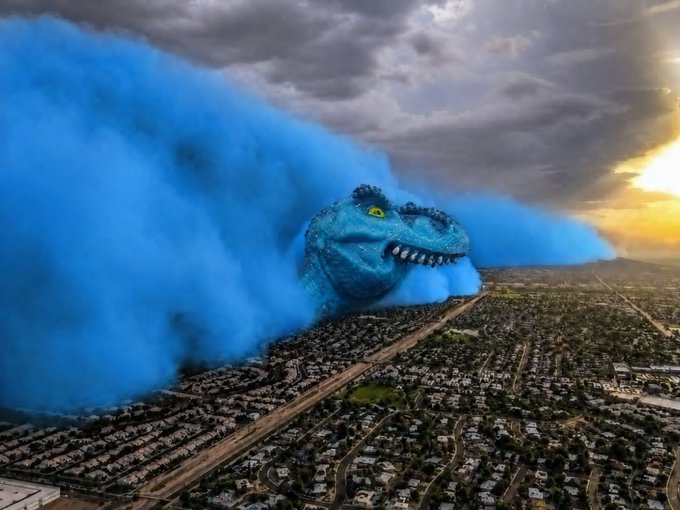 A surreal aerial view shows a giant blue T-Rex head emerging from blue smoke clouds over a sprawling suburban neighborhood at sunset.