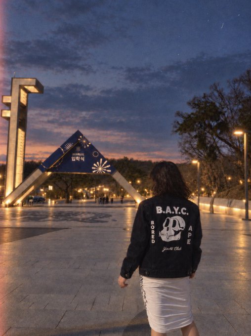 A woman wearing a black "B.A.Y.C. BORED APE YACHT CLUB" jacket and white skirt stands facing an entrance archway marked "2023" and Korean text at dusk. The BAYC-branded apparel ties directly into the post's "B.A.Y.C." reference, showcasing streetwear associated with the popular NFT collection.