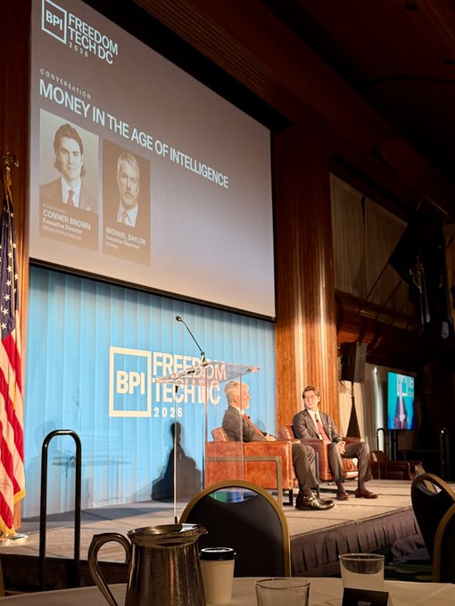 Two men sit on stage at the 'BPI FREEDOM TECH DC 2026' convention during a panel titled 'MONEY IN THE AGE OF INTELLIGENCE' featuring Conner Brown and Michael Saylor.
