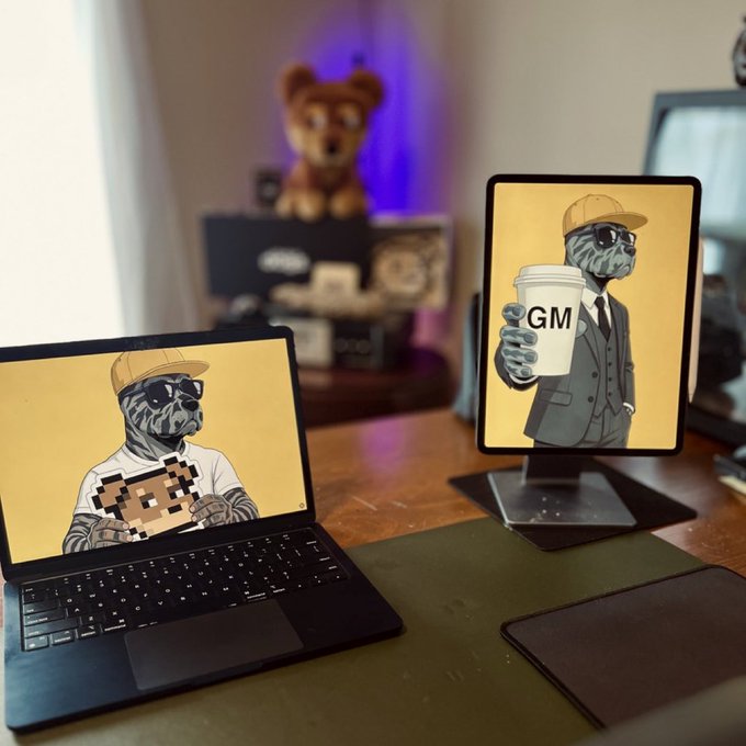 A desk setup featuring a laptop and monitor both displaying artwork of a dog in sunglasses and a yellow cap, with one holding a 'GM' coffee cup; a teddy bear sits in the background.