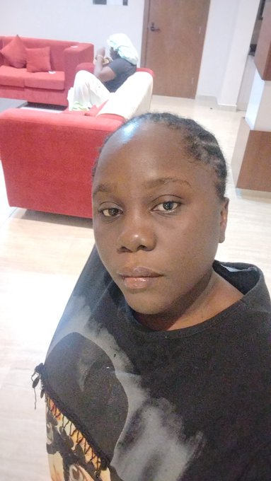 A selfie of a young woman with a shaved head wearing a black t-shirt, posing in a living room with red furniture in the background.