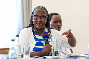 A woman with glasses and braided hair wearing a white cardigan smiles and points while holding a microphone during what appears to be a discussion or panel.