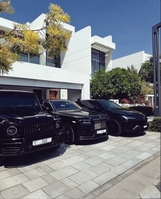 Three black luxury SUVs, including a Mercedes G-Wagon and a Rolls-Royce, are parked in the driveway of a white modern villa.