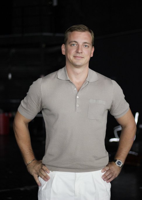 A man with short blond hair wearing a beige polo shirt and white pants stands with his hands on his hips in a dimly lit setting.