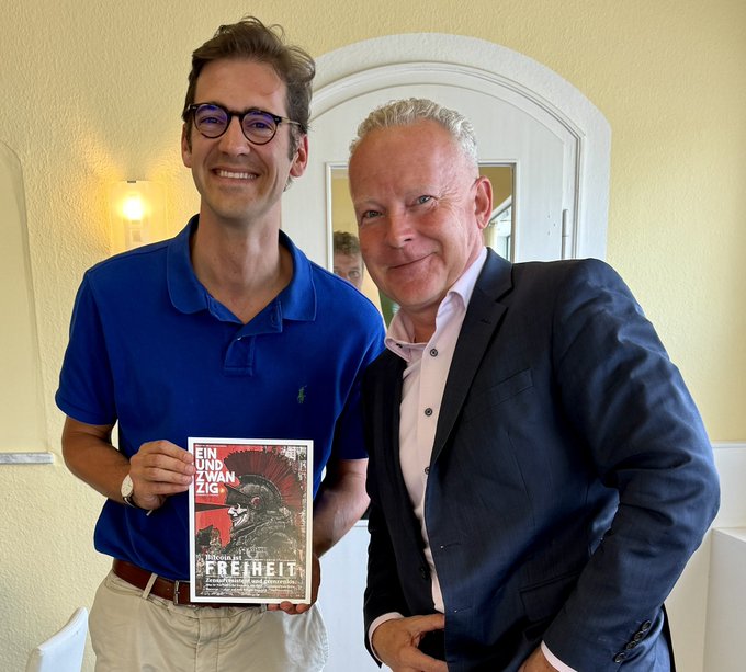 Two men smile together indoors, with one holding a book titled "Einundzwanzig: Warum wir Freiheit brauchen" featuring an image of a Spartan helmet. The photo accompanies a post about meeting with Dr. Friedrich Dorn, the CDU/CSU parliamentary group's expert on Bitcoin, arranged through Bundestag representative Dr. Pilsinger.
