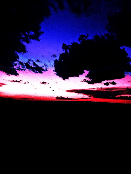 A dark landscape photograph showing a dramatic sunset or sunrise with dark clouds, a deep blue sky, and a vibrant red horizon line above a silhouetted ground.