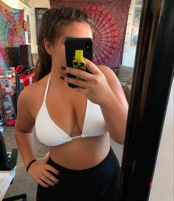 A woman in a white bikini top and black bottoms takes a mirror selfie in a room with tapestries and a vacuum visible in the background, while the phone case displays the green "X" Stake.com logo—tied to the post's promotion of becoming a "Stake VIP Manager."