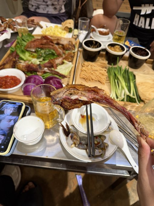 A person holds up a partially eaten large rib bone over a tray of assorted dishes including meats, noodles, vegetables, and a teapot, with another diner wearing a shirt that reads "HALL" visible across the table. The lavish spread of food and drinks illustrates the caption's message about indulging freely—seemingly without immediate financial concern—and putting the meal "on credit."