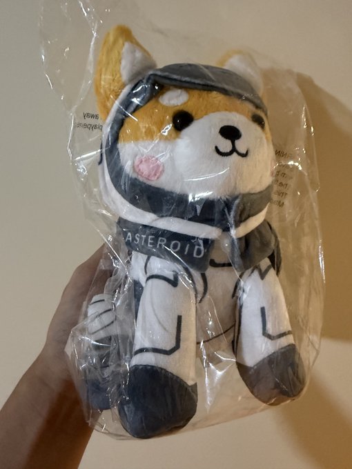 A hand holds a Shiba Inu plush toy dressed in an astronaut suit labeled 'ASTEROID', packaged in clear plastic.