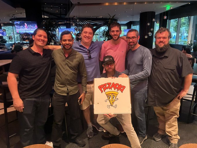 A group of seven men pose together in a modern office lounge, with one man kneeling to hold up a canvas painted with 'PIZZACORN', a cartoon pizza slice with a unicorn horn.