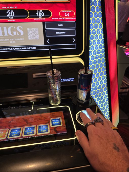 A hand wearing rings rests on a slot machine console at what appears to be a casino, where two cans of Red Bull are propped up beside the touchscreen—directly supporting the post's claim that casinos will freely provide complimentary energy drinks to players. The screen displays "HOLIDAY GIFT SHOPPE" and a balance of "1¢" credits won, linking to the @RareEvo question about whether the event will offer similar complimentary beverages.