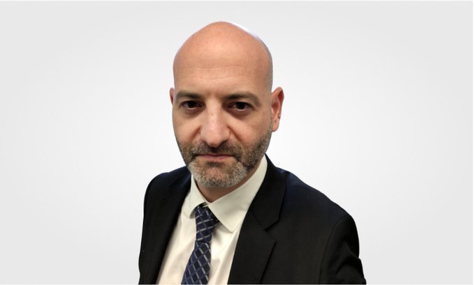 A professional headshot of a bald, bearded man in a dark suit and patterned blue tie, photographed against a plain light gray background.
