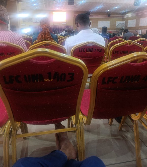 A view from the back of a hall with gold-framed chairs labeled 'LFC UWA 1403' and similar, facing a stage where people are seated.