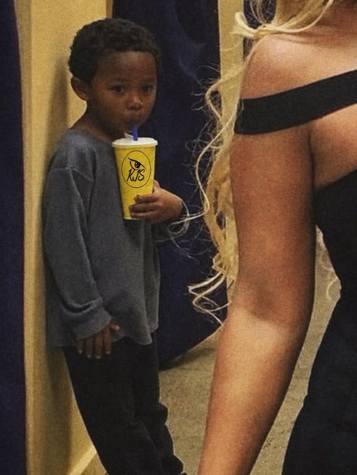 A young boy in a gray long-sleeved shirt and dark pants stands against a wall sipping from a yellow 'Rally's' cup, next to a blonde woman in a black off-shoulder dress.