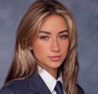A portrait of a young woman with long blonde hair wearing a dark blazer, white shirt, and tie against a blue backdrop.