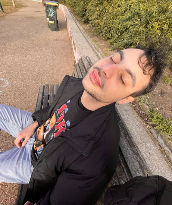 A young man with curly dark hair and a light beard wearing a black jacket over a graphic t-shirt, reclining peacefully on a park bench with his eyes closed.