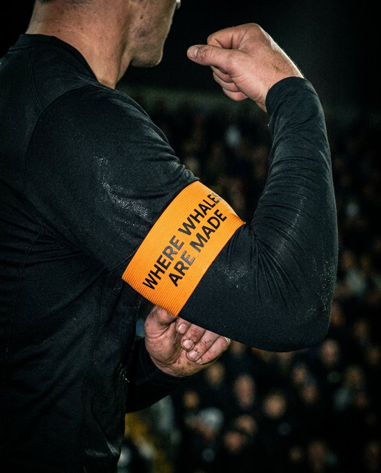 A muscular person in a black long-sleeve shirt flexes their arm, displaying a bright orange armband reading "WHERE WHALES ARE MADE" against a blurred crowd backdrop. The image ties into the "Made for the Whales" theme by visually emphasizing the message on the armband, reinforcing the idea that strength and commitment are forged in this environment.