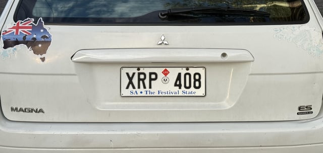 The rear of a white Mitsubishi Magna ES features a South Australian license plate reading "XRP 408" with the slogan "SA – The Festival State," playfully interpreted by the poster as a crypto ticker sign from above.
