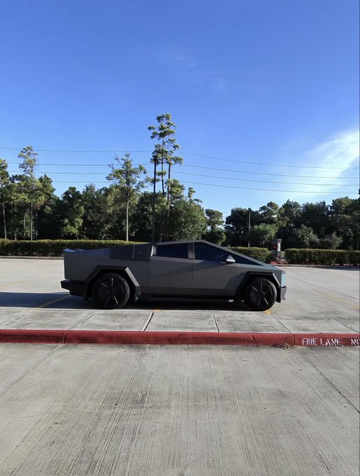 A dark gray Tesla Cybertruck is parked in a lot under a clear blue sky, with 'FIRE LANE NO' painted on the curb.
