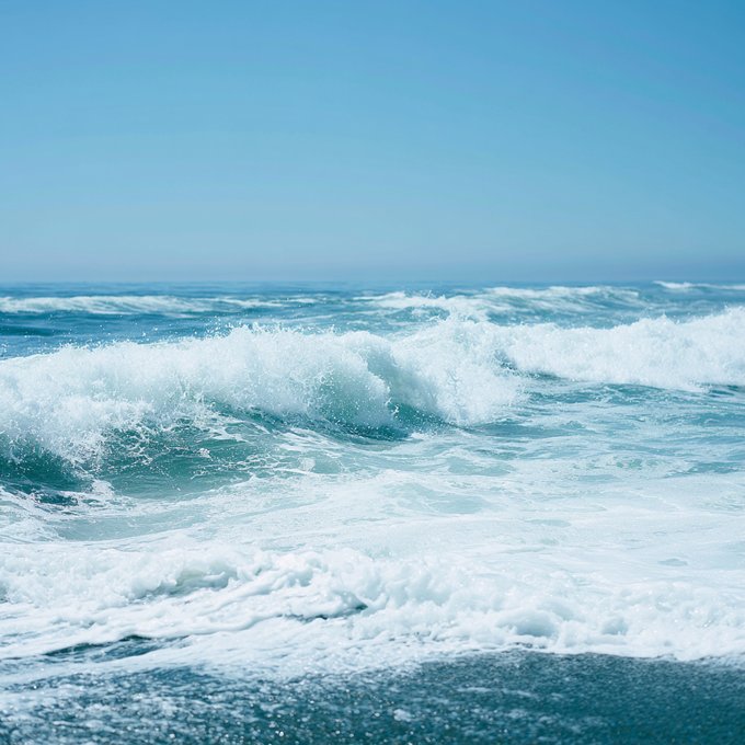 A dynamic ocean wave crashes and foams against the shoreline under a clear blue sky, its rolling cresting waves resembling a "gm" (good morning) wave greeting the day.