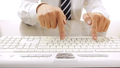 A person in business attire aggressively jabs at a keyboard with both index fingers, humorously illustrating the joke about using overly forceful or curt typing style when communicating with AI chatbots versus colleagues.
