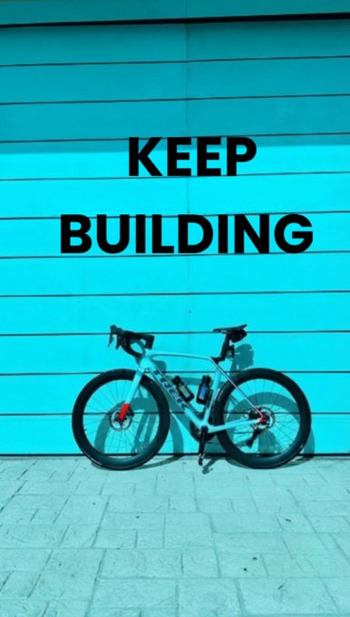 A photograph of a Trek bicycle leaning against a bright turquoise wooden wall featuring the bold black text 'KEEP BUILDING'.