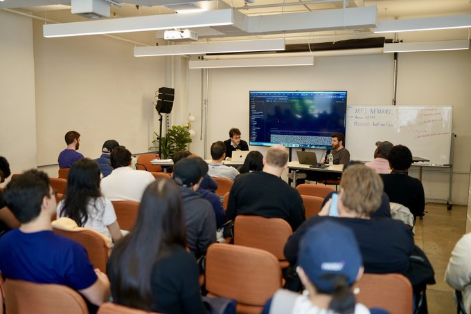 A classroom or seminar setting where a group of attendees sits in chairs facing a large screen displaying code, while a presenter stands near the front beside a whiteboard with handwritten notes.