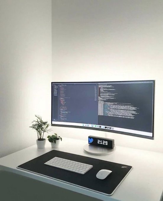 A clean minimalist white desk setup features a curved monitor displaying code, a wireless keyboard and mouse on a large mousepad, small potted plants, and a digital clock showing '21:29'.